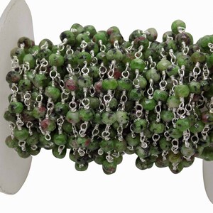 May include: A strand of green and maroon gemstone beads, strung on a silver chain, is coiled on a white spool. The beads are round and faceted, with a mix of green and maroon colors. The silver chain adds a metallic contrast.