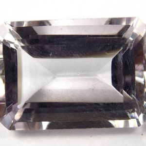 Puede incluir: Una gema rectangular transparente con un diseño de talla escalonada. La gema tiene una apariencia transparente con múltiples facetas que reflejan la luz. La forma es un corte esmeralda clásico, que muestra la claridad y el brillo de la piedra.