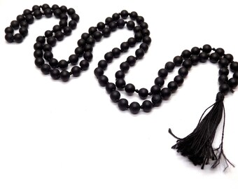 Puede incluir: Un collar de cuentas negro con una borla. El collar está hecho de cuentas negras mate ensartadas. La borla está hecha de hilos negros. El collar se muestra sobre un fondo blanco.