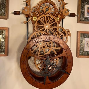 May include: A wooden wall clock with exposed gears and Roman numeral hour markers. The clock has a dark brown finish with intricate wooden gears and a black clock hand. The clock is mounted on a wall with framed artwork.