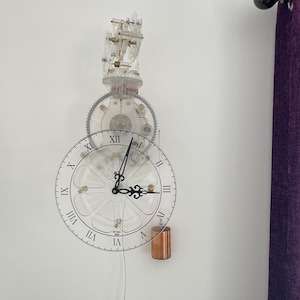 May include: A clear, transparent wall clock with visible gears and a pendulum. The clock face has black Roman numerals and hands. A copper-colored cylinder hangs below. The clock is mounted on a white wall.