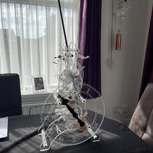 May include: A clear acrylic clock with a black pendulum and gold gears. The clock face has Roman numerals and is set on a black table.