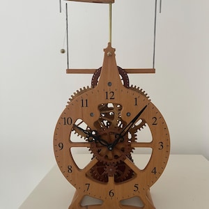 Peut inclure: Une horloge en bois avec des engrenages visibles et un pendule. Le cadran est circulaire avec des chiffres romains. L'horloge est en bois brun clair et a un design unique. Le pendule oscille. L'horloge mesure environ 30 cm de haut.
