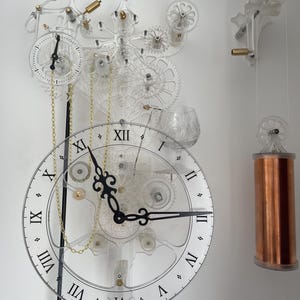 Pode incluir: Um relógio de acrílico transparente com um mostrador preto e marcadores de hora em algarismos romanos. O relógio tem um mecanismo complexo com engrenagens e correntes. O relógio está pendurado em uma parede branca com um peso de cobre pendurado do lado.