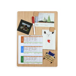Puede incluir: Un tablero de corcho de madera con una variedad de artículos fijados a él, incluyendo una foto de dos niños, un dibujo de un oso, un horario, una foto de árboles y una pequeña silla de cerámica. El tablero de corcho es de color marrón claro y tiene una veta de madera natural.