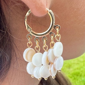 May include: Gold-tone hoop earrings with dangling white and cream-colored shell discs. The earrings feature a circular hoop design with multiple chains suspending the shell accents. The shell discs have a natural, organic appearance.
