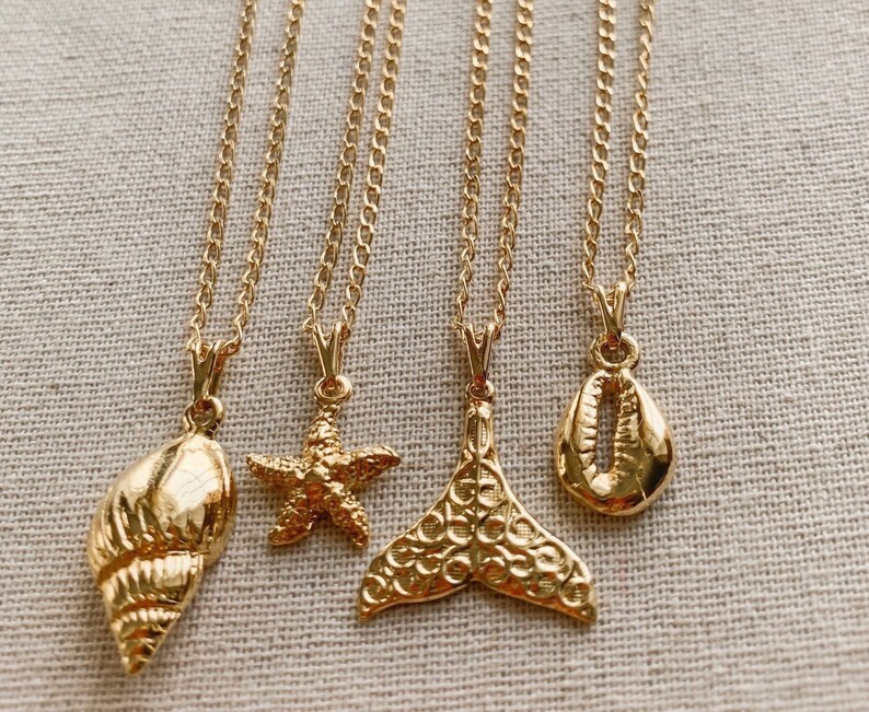 May include: Four gold-colored necklaces with ocean-themed pendants. The pendants include a seashell, starfish, mermaid tail, and a cowrie shell. Each necklace has a delicate chain and a lobster clasp.