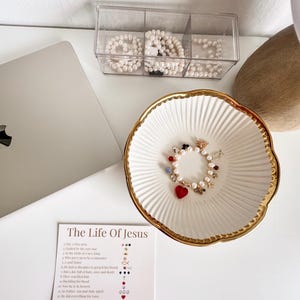 May include: A white ceramic bowl with a gold scalloped rim containing a pearl charm bracelet with red heart, star, and fish accents. Beside it is a card titled The Life Of Jesus, detailing twelve religious symbols and their meanings in a list format.