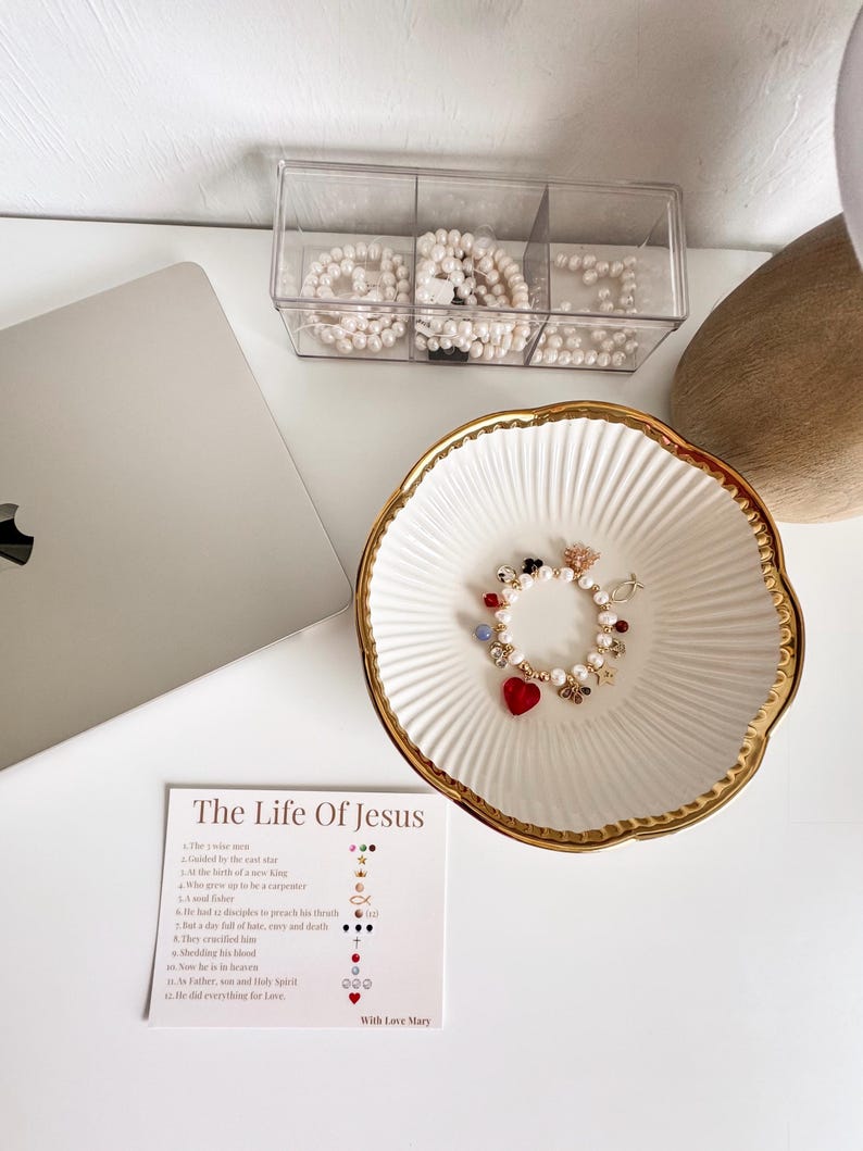 May include: A white ceramic bowl with a gold scalloped rim containing a pearl charm bracelet with red heart, star, and fish accents. Beside it is a card titled The Life Of Jesus, detailing twelve religious symbols and their meanings in a list format.