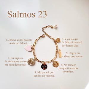 Puede incluir: Pulsera dorada con dijes variados, incluyendo un coraz&oacute;n, una estrella y un sol. La pulsera tiene una perla al final y texto en espa&ntilde;ol: "Salmos 23". Los dijes est&aacute;n detallados con dise&ntilde;os intrincados y texto.