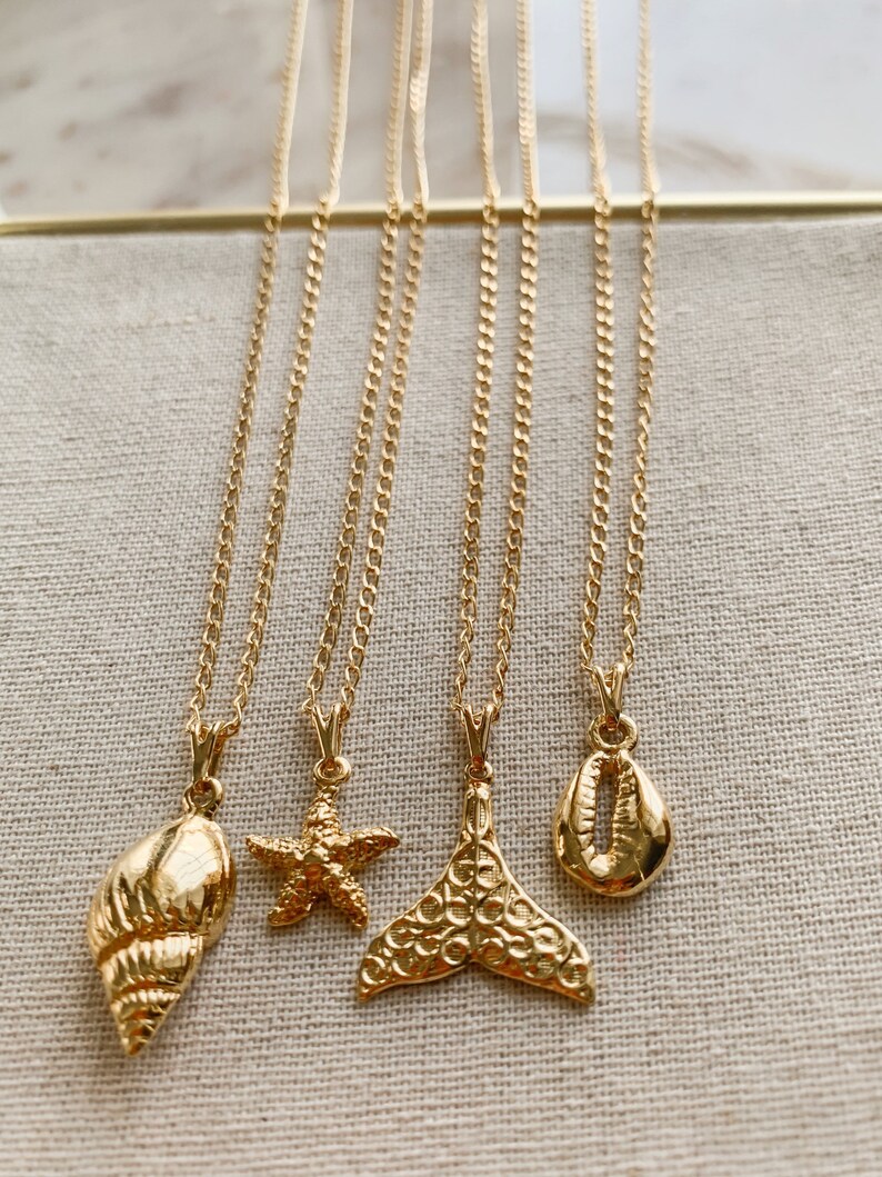 May include: Five gold-colored necklaces with different pendants: a seashell, a starfish, a mermaid tail, and a cowrie shell. The necklaces hang against a neutral-colored fabric background. The chains are delicate and the pendants are detailed.