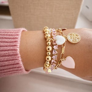 Puede incluir: Pulseras doradas con varios dijes y cuentas. Una pulsera presenta cuentas redondas doradas, otra tiene piedras rosas con forma de cristal y una tercera tiene una cadena con un dije de coraz&oacute;n blanco, un dije de coraz&oacute;n rosa y un dije de moneda dorada.