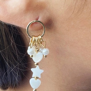 May include: Gold-tone dangle earrings with a circular top and multiple charms. The charms include white heart and star shapes, and small white beads. The earrings are designed to be worn in the ear.