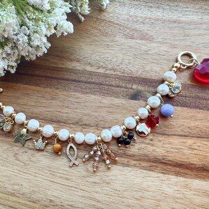 May include: A white pearl bracelet with gold charms, including a crown, a fish, a star, and a heart. The bracelet has a toggle clasp and is adorned with red, blue, and green gemstones.