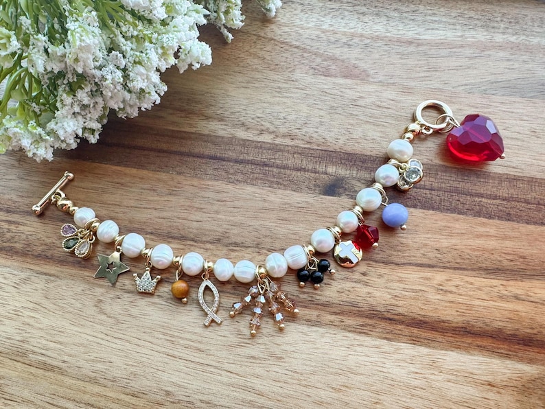 May include: A white pearl bracelet with gold charms, including a crown, a fish, a star, and a heart. The bracelet has a toggle clasp and is adorned with red, blue, and green gemstones.