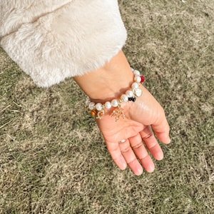 May include: A white pearl bracelet with gold charms and colorful beads. The bracelet is on a person's wrist.
