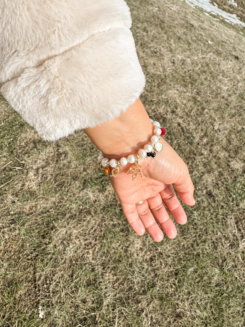 May include: A white pearl bracelet with gold charms and colorful beads. The bracelet is on a person's wrist.