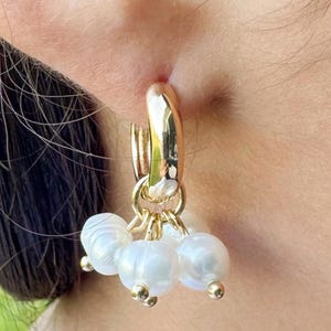 May include: Gold hoop earrings with three dangling white pearl clusters. The earrings have a polished finish and are shown against a blurred background.