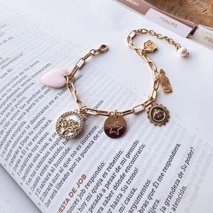 Puede incluir: Pulsera dorada con dijes que incluye un coraz&oacute;n rosa, una estrella, un &aacute;rbol de la vida, un sol, una botella y una perla. La pulsera se muestra sobre un libro abierto con texto.