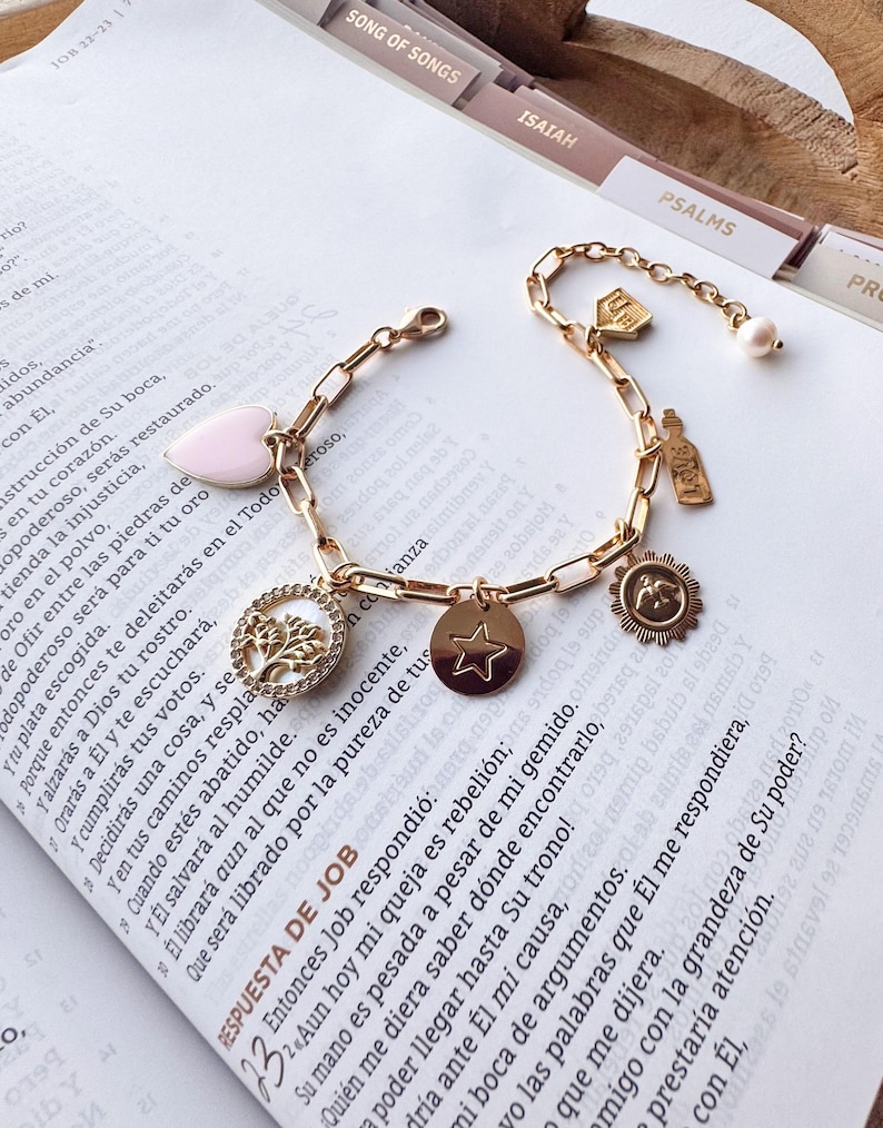 Puede incluir: Pulsera dorada con dijes que incluye un coraz&oacute;n rosa, una estrella, un &aacute;rbol de la vida, un sol, una botella y una perla. La pulsera se muestra sobre un libro abierto con texto.