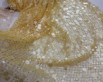 May include: A roll of pale yellow fabric with a net-like weave, heavily embellished with large, round, iridescent sequins. The fabric is partially unrolled, revealing its texture and the way the sequins catch the light.