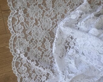 May include: White floral lace fabric with a scalloped edge. The lace features a detailed pattern of flowers and leaves. The fabric is draped, showing the texture and design. Ideal for sewing projects, bridal wear, or home decor.