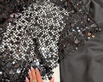 May include: Black sequin fabric with a net backing. The sequins are round and shiny, creating a textured surface. The fabric is draped over a black satin material. A hand with a gold ring is visible.