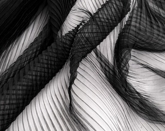 May include: Close-up of a black and white pleated fabric. The fabric has a striped pattern with sections of woven, textured detailing. The folds create shadows and depth, highlighting the contrast between the black and white elements. The material appears to be lightweight and flowing.
