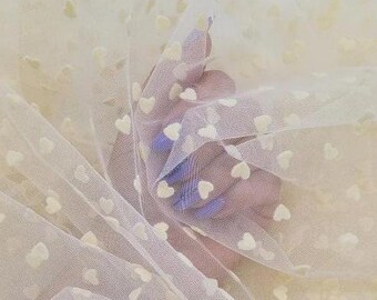 May include: Close-up of a sheer, off-white fabric with a delicate heart pattern. The fabric is gathered, creating soft folds and shadows. The hearts are small and evenly spaced across the material.