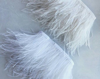 May include: Two white ostrich feather trims, each with a white fabric backing. The trims are soft and fluffy, with long, delicate feathers.