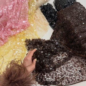 May include: A collection of sequined fabric in various colors, including pink, yellow, beige, black, and brown. The fabrics are arranged in a layered display, showcasing the texture and sheen of the sequins. The image highlights the different colors and textures of the fabric.