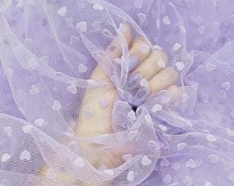 May include: A close-up of a sheer, lavender fabric with a delicate heart pattern. The fabric is gathered in the center, creating soft folds and shadows. The hearts are small and evenly spaced across the material.