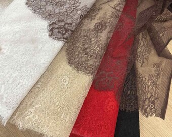 May include: A collection of folded lace fabrics in various colors, including white, beige, red, brown, and black. The lace features intricate floral patterns and scalloped edges, creating a delicate and textured appearance. The fabrics are layered, showcasing the different colors and designs.