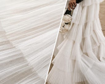 May include: A floor-length white dress featuring multiple horizontal tiers of pleated tulle fabric. The skirt is constructed with overlapping layers of sheer, fine-mesh material that create a voluminous, textured appearance throughout the garment.