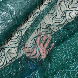 May include: Close-up of emerald green fabric with a shimmering, glittery overlay. The fabric features a delicate, swirling pattern in darker green, creating a textured effect. The material appears to be sheer, with a hand visible beneath.