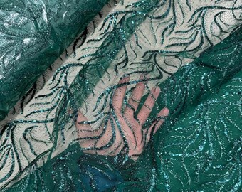 May include: Close-up of emerald green fabric with a shimmering, glittery overlay. The fabric features a delicate, swirling pattern in darker green, creating a textured effect. The material appears to be sheer, with a hand visible beneath.