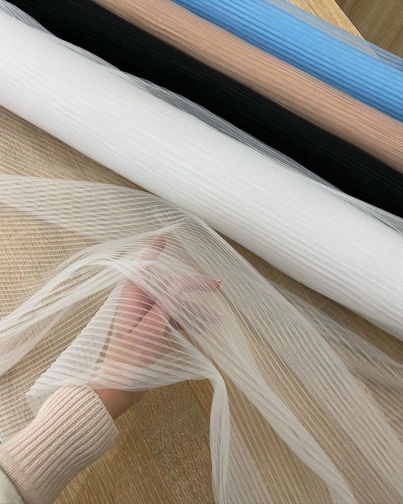 May include: White, black, light blue, and beige striped tulle fabric rolls. The fabric is sheer and has a delicate texture.
