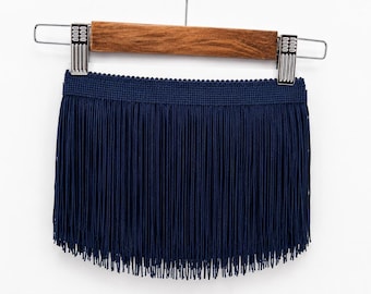 May include: Navy blue fringe trim, displayed on a wooden hanger with metal clips. The trim features a woven band at the top and long, straight fringe. The fringe is a solid, dark blue color, and the trim is against a white background.