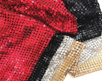 May include: Close-up of a collection of sequined fabrics in red, black, silver, and gold. The sequins are sewn onto a mesh backing, creating a shimmering effect. The fabrics are folded and layered, showcasing the different colors and textures.