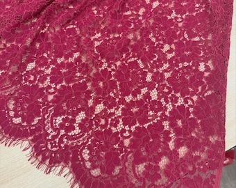 May include: Close-up of a burgundy lace fabric with a floral pattern. The delicate lace is draped, revealing a glimpse of a lighter background. The edges of the fabric are scalloped, adding a decorative touch to the textile.