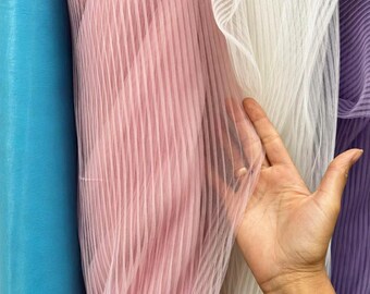 May include: Close-up of colorful fabric rolls. The image features rolls of sheer fabric in shades of blue, pink, white, and purple. The fabric has a vertical striped texture. A hand is touching the white fabric.