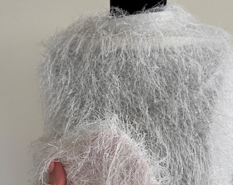 May include: A white, fluffy, textured garment, possibly a sweater or top, with long, thin strands creating a soft, shimmering effect. The fabric appears to be held by a hand, showcasing its delicate, airy quality. The garment is displayed on a black mannequin.