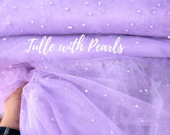 May include: A swatch of lavender tulle fabric with scattered pearl embellishments. The fabric is folded and draped, with the text "Tulle with Pearls" in white script across the front. The fabric is soft and translucent.