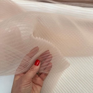 May include: Pale pink tulle fabric with a delicate grid pattern. The fabric is pleated and has a soft, flowing texture.