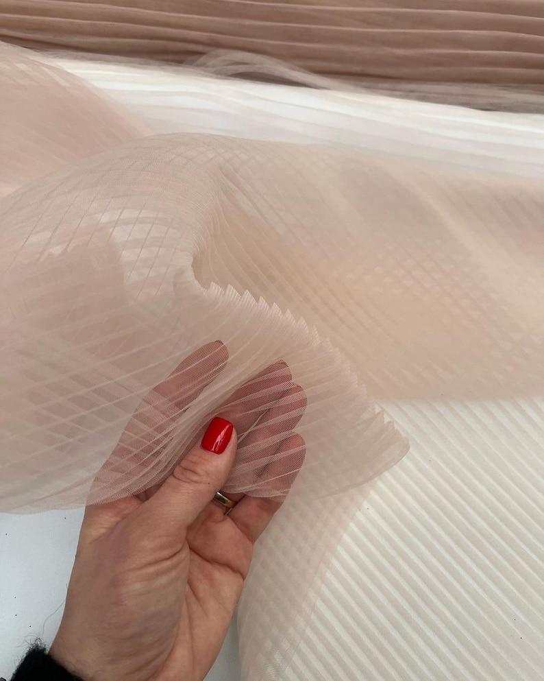 May include: Pale pink tulle fabric with a delicate grid pattern. The fabric is pleated and has a soft, flowing texture.