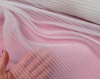 May include: Close-up of white and pink pleated fabric. The fabric has a sheer, layered appearance with vertical pleats. The pink color transitions from a darker shade at the bottom to a lighter shade at the top. A hand is visible, touching the fabric.