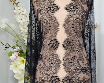 May include: Black lace fabric with a floral pattern is draped over a mannequin. The fabric is sheer, revealing the mannequin's form. The lace features intricate floral designs and a scalloped edge at the neckline. The background includes white flowers.