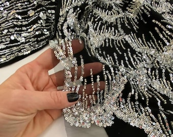 May include: Black fabric heavily embellished with silver sequins and beads. The design features intricate patterns of sequins and beaded fringe. The fabric appears to be part of a garment, possibly a dress or top, with a sheer overlay.
