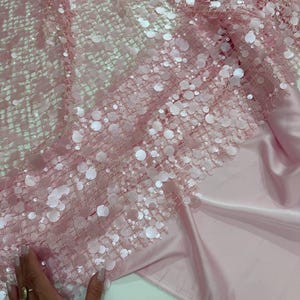 May include: Pink fabric with a sequined overlay. The fabric has a net-like base with large, round sequins scattered across the surface. The sequins are a lighter shade of pink, creating a shimmering effect. The fabric is draped over a smooth, satin-like material.
