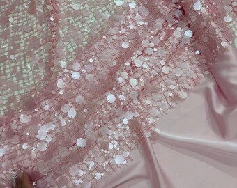 May include: Pink fabric with a sequined overlay. The fabric has a net-like base with large, round sequins scattered across the surface. The sequins are a lighter shade of pink, creating a shimmering effect. The fabric is draped over a smooth, satin-like material.